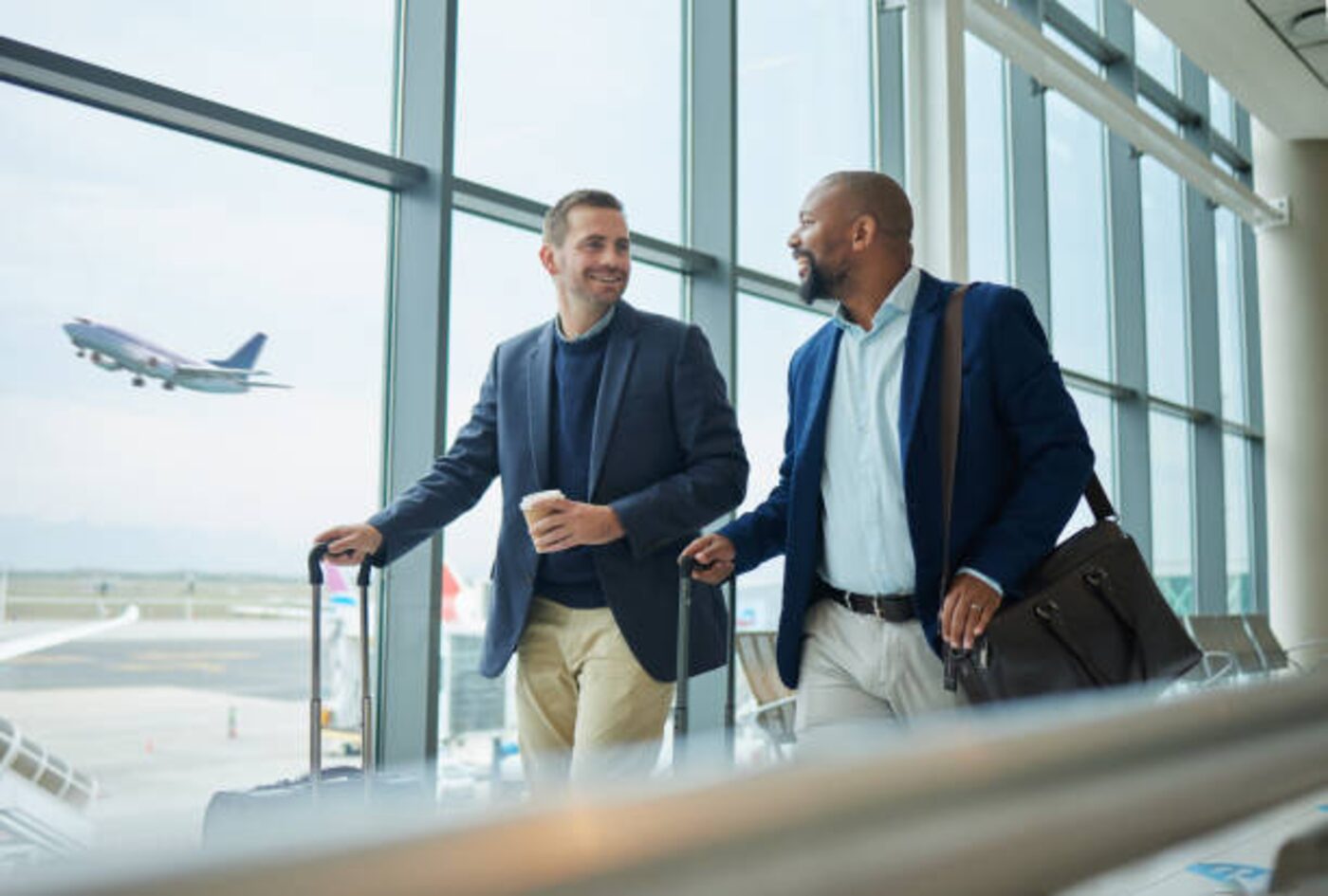 Two colleagues with their luggage at an airport window, an aircraft climbing behind them.